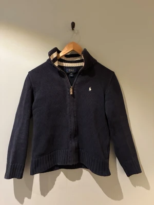 Marinblå ziptröja från Polo Ralph Lauren - Mörkblå stickad ziptröja från Polo Ralph Lauren med krage och har även en dragkedja framtill och en liten vit logga broderad på bröstet. Insidan av kragen har randigt mönster i vit och marinblått. Perfekt för dig som är 150-155 cm.
