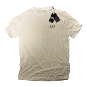 Vit EA7 Emporio Armani t-shirt herr - Snygg vit t-shirt i nyskick med prislapp från EA7 Emporio Armani med diskret logga på bröstet. Klassisk rund halsringning och korta ärmar. Tillverkad i mjuk bomull för en bekväm känsla och stilren look. Tröjan är i storlek L herr