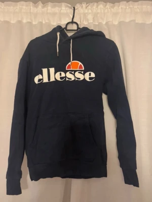 Mörkblå Ellesse hoodie med logga - Snygg mörkblå hoodie från Ellesse med stor vit logga och orange detalj på bröstet. Klassisk känguruficka framtill, huva med vita snören och mjukt material på insidan. Perfekt för en avslappnad och sportig stil.