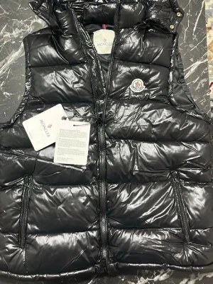 Svart dunväst från Moncler - Snygg svart dunväst från Moncler med glansig finish, hög krage och dragkedja framtill. Västen har två stora fickor med dragkedja och Moncler-logga på bröstet. Insidan har ett unikt serietidningsmönster och etiketter med info om material och skötsel.