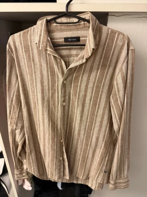 Beige och vit randig linneskjorta - Säljer en beige och vit randig skjorta från Minimum i luftigt linne. Skjortan har klassisk krage, knappar framtill och långa ärmar. Perfekt för en avslappnad men snygg stil.