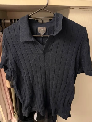Mörkblå ribbad polotopp från H&M - Mörkblå ribbad polotopp från H&M med korta ärmar och krage. Toppen har en relaxed fit och är tillverkad i mjukt material som känns skönt mot huden. Perfekt till jeans eller kjol för en enkel men snygg look.