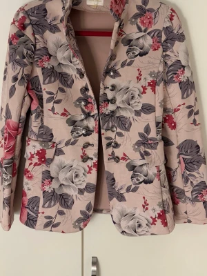 Blommig kavaj i rosa och grå toner - Snygg kavaj från Chica Londonmed blommigt mönster i rosa, grått och vitt. Kavajen har flera dekorativa knappar framtill, två fickor och en figurnära passform. Materialet känns mjukt och har en lätt glans. Perfekt för dig som vill sticka ut med din outfit.