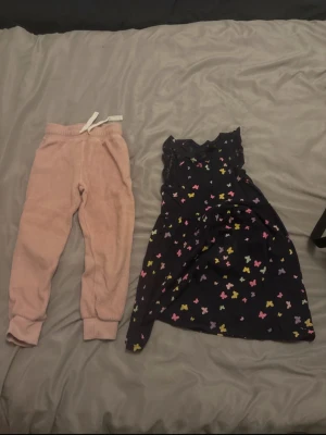 Ljusrosa mjukisbyxor med dragsko - Mysiga ljusrosa mjukisbyxor med resår i midjan och vit dragsko. Tillverkade i ett mjukt fleecematerial som känns skönt mot huden. Perfekta för chill dagar hemma eller när du vill vara extra bekväm. Klassisk passform med muddar vid bensluten.  Storlek på byxorna: 110-116 och på klänningen är det 4-6 år.