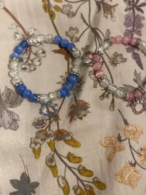 Två pärlarmband med bokstavsberlocker - Säljer två elastiska armband med pärlor i blått och rosa, kombinerat med genomskinliga glaspärlor och silverfärgade detaljer. Ena armbandet har en blå bokstavsberlock och det andra en rosa bokstavsberlock. Perfekta för att ge din stil en personlig touch.