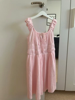Ljusrosa sommarklänning Annie S - Söt ljusrosa klänning från Annie i storlek S. Klänningen har breda axelband med volangdetaljer, smockad överdel och en liten knytning framtill. Tillverkad i ett lätt, skrynkligt tyg som passar perfekt för varma dagar.