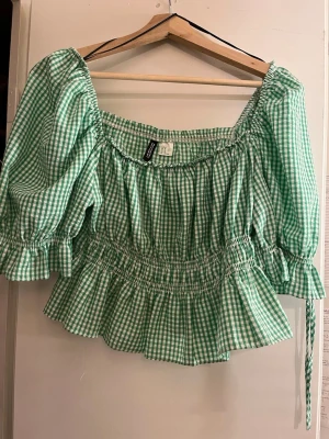 Grön rutig blus från H&M Divided - Supersöt grön och vit rutig blus från H&M Divided med puffärmar med knytning, volangkant och smock i midjan. Blusen har en fyrkantig ringning.