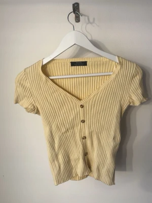 Ribbad gul topp från Bershka - Kortärmad ribbad topp från Bershka i ljusgul färg. Toppen har v-ringning, knappar framtill och en figurnära passform. Perfekt till jeans eller kjol för en trendig look.