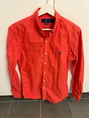 Röd skjorta från Ralph Lauren, stl S - Klassisk röd skjorta från Ralph Lauren med button-down krage och blå broderad logga på bröstet. Skjortan har långa ärmar, vita knappar och är tillverkad i bomull. Perfekt för en stilren och avslappnad look.