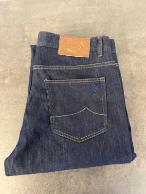 Mörkblå raka jeans från Jacob Cohen, stl 34 - Snygga mörkblå jeans från Jacob Cohen med klassisk femficksmodell och raka ben. Jeansen har kontrastsömmar, läderpatch bak i midjan och är tillverkade i slitstark denim. Perfekta för en avslappnad och stilren look.