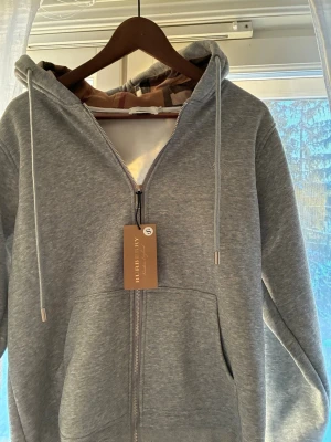 Grå zip hoodie från Burberry - Snygg grå hoodie från Burberry med dragkedja, huva med dragsko och klassisk Burberry-logga broderad på bröstet. Hoodien har känguruficka framtill och ribbade muddar. Tillverkad i mjukt bomullsmaterial för extra komfort.