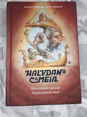 Halvdan & Meia: Hövdingens bägare - En samlingsvolym med två spännande vikingaäventyr av Martin Widmark och Mats Vänehem. Boken har hårda pärmar i rödbrun ton med illustrerat omslag som visar vikingar, skepp och äventyrsscener. Perfekt för dig som gillar historiska berättelser och äventyr.