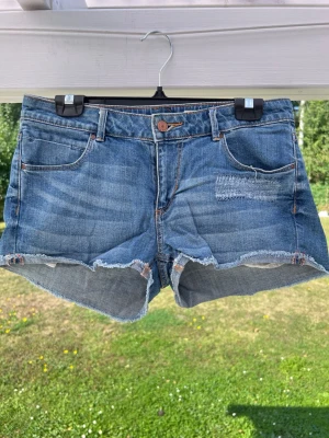 Y2k Lågmidjade jeansshorts med rå kant - Snygga blå jeansshorts med rå, fransig kant och klassisk femficksdesign. Storlek 40 och har en slitning på vänster ben. Midja: 86 cm 