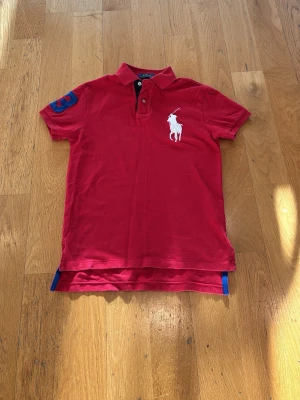 Röd pikétröja från Ralph Lauren - Klassisk röd pikétröja från Ralph Lauren med stor vit logga på bröstet och blå detalj på ärmen. Tröjan har krage, knappar vid halsen och korta ärmar. Perfekt för en stilren och sportig look.
