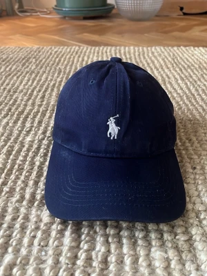 Mörkblå keps från Polo Ralph Lauren - Säljer en marin blå keps från polo Ralph lauren, köpt på nätet för typ 1 år sedan men har ledsnat på den. Den är använd men ändå i fint skick. No size