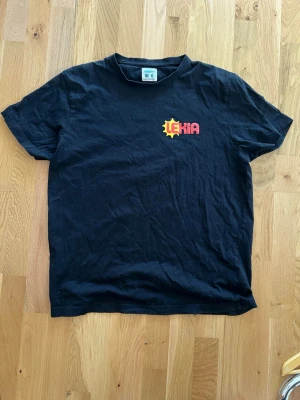Svart Lekia t-shirt i bomull - Svart t-shirt från Lekia med tryckt logga i gult och rött på bröstet. Klassisk rak passform och rund halsringning. Tillverkad i mjuk bomull, perfekt för en avslappnad och enkel stil. L men sitter som M
