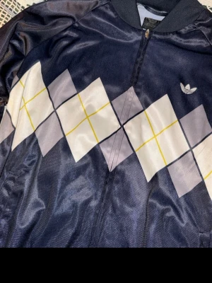 Mörkblå vindjacka från Adidas M - Snygg mörkblå vindjacka från Adidas med dragkedja och klassiskt argyle-mönster i vitt, grått och gult på framsidan. Jackan har ribbad krage och Adidas-logga på bröstet. Perfekt för dig som gillar sportig retrostil.