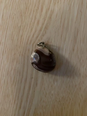 Brunt och vitt stenberlock halsband - Unik halsbandsberlock i brunt och vitt med naturligt mönster. Berlocken har en mjukt rundad form och är tillverkad av polerad sten med ett metallfäste för kedja. Perfekt för dig som gillar naturliga material och jordnära färger.