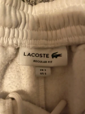 Vita mjukisbyxor från Lacoste - Säljer ett par vita mjukisbyxor från Lacoste med regular fit. Byxorna har resår i midjan och dragsko för skön passform. Perfekta för chill dagar eller när du vill ha det extra bekvämt. Klassisk Lacoste-logga på etiketten.