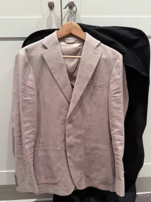 Beige linnékavaj från John Henric - Snygg beige kavaj i linne med klassisk passform och två knappar framtill. Kavajen har två stora framfickor, bröstficka och slits bak. Perfekt för en stilren och avslappnad look. Passar dig mellan 170-180