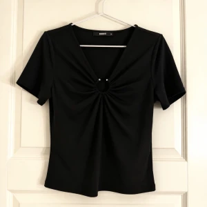 Svart topp med ringdetalj från Bik Bok - Snygg svart topp från Bik Bok med v-ringning och en dekorativ ringdetalj framtill. Kortärmad modell i mjukt stretchigt material som sitter figurnära. Användt några enstaka gånger.