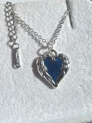 Halsband med blått hjärthänge i stål - Lyser i mörker - Snyggt halsband i silverfärgat rostfritt stål med ett blått hjärthänge inramat av vingar. Kedjan är tunn och har förlängningslänk. Perfekt accessoar för dig som gillar unika detaljer och cool design. Hänget lyser i mörker efter att ha varit en kort stund i vanligt dagsljus och lyser vanligtvis i flera timmar. 