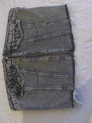 Grå denim tubtopp med dragkedja - Cool tubtopp i grå denim med råa kanter och dragkedja framtill. Toppen har dekorativa fickor med knappar och fransiga detaljer längs nederkanten. Perfekt för dig som gillar edgy och unik stil.