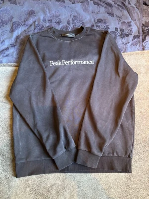 Svart sweatshirt från Peak Performance - Mörkgrå sweatshirt från Peak Performance med klassisk rund hals och vit logotyp på bröstet. Tröjan har ribbade muddar vid ärmslut och nederkant. Perfekt för en avslappnad och stilren look.