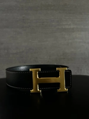 Svart Herme’ss bälte  - Stilrent svart skärp från Herm’es med ett stort H-format spänne i guld. Skärpet är tillverkat i skinn och har vita kontrastsömmar längs kanterna. Perfekt för att lyfta din outfit med en lyxig touch.