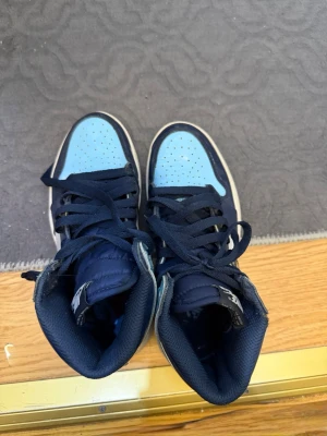 Nike Air Jordan 1 High blå/vit - Snygga Nike Air Jordan 1 High sneakers i blått och vitt med mörkblå detaljer och klassisk swoosh på sidan. Skorna har högt skaft, perforerad tåbox och är tillverkade i en mix av läder och syntetmaterial. Perfekta för dig som vill sticka ut med din stil. Priset går att diskutera 