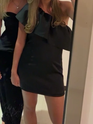 Svart one shoulder kortklänning - Snygg svart kortklänning med one shoulder-design och stor volangdetalj över axeln. 