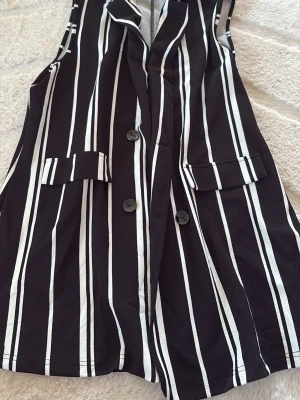 Svartvit randig kortklänning - Snygg svart kortklänning med vita vertikala ränder och knappar framtill. Klänningen har fickdetaljer och en stilren, modern look. Perfekt för dig som gillar trendiga och grafiska mönster.