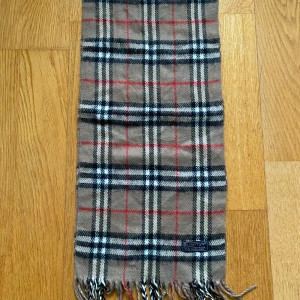 Beige rutig halsduk från Burberrys - Klassisk beige halsduk från Burberrys i 100% lammull. Den har det ikoniska rutmönstret i beige, svart, vitt och rött samt fransar i ändarna. Perfekt accessoar för att lyfta din outfit med en tidlös brittisk touch.