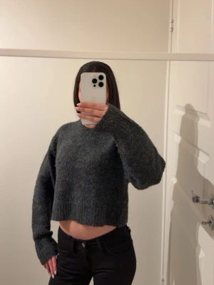 Mörkgrå croppad stickad tröja - Mysig mörkgrå stickad tröja med croppad passform och långa ärmar. Tröjan har en enkel design med rund halsringning och ribbstickade muddar. Perfekt för lager på lager och en avslappnad look.