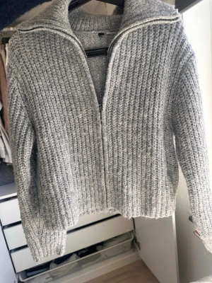 Grå stickad ziptröja med krage - Mysig grå stickad tröja med dragkedja framtill och hög krage. Tröjan har en grovstickad struktur och långa ärmar, perfekt för kyliga dagar. Passar till både jeans och kjol för en avslappnad look.
