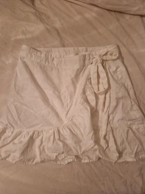 Vit omlottkjol  - Söt vit kort omlottkjol från NLY Trend med volangkant och knytband i midjan. Kjolen är gjord i ett lätt bomullstyg och har en feminin och somrig look.