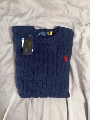 Marinblå kabelstickad tröja Polo Ralph Lauren - Kabelstickad marinblå tröja från Polo Ralph Lauren med rund halsringning och den klassiska röda logotypen broderad på bröstet. Tröjan har långa ärmar och ribbade muddar vid ärmslut och nederkant. Perfekt för dig som gillar stilren och tidlös design. mått: axelbredd 40cm. längd 64cm.