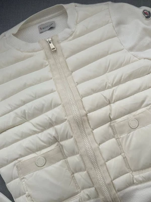 Moncler cardigan dam - Snygg vit dunväst från Moncler med quiltad design och två framfickor med tryckknappar. Västen har dragkedja framtill och ribbade detaljer vid kanterna. Klassisk Moncler-logga på axeln och stilren look som passar till många outfits.