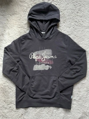 Mörkgrå hoodie från Pepe Jeans - Snygg mörkgrå hoodie från Pepe Jeans med tryckt logga och texten 'London' på bröstet. Hoodien har en klassisk känguruficka framtill och ribbade muddar vid ärmslut och nederkant. Tröjan är i storlek 164cm och i mycket bra skick✅ Fraktar inom en dag✅ Priset är inte hugget i sten🙂
