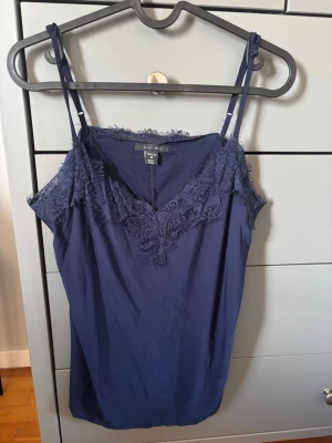 Mörkblå linnetopp med spets - Snygg mörkblå linnetopp från Amisu med tunna axelband och vacker spetsdetalj längs ringningen. Perfekt att styla till jeans eller kjol för en trendig look. Materialet känns lätt och svalt.