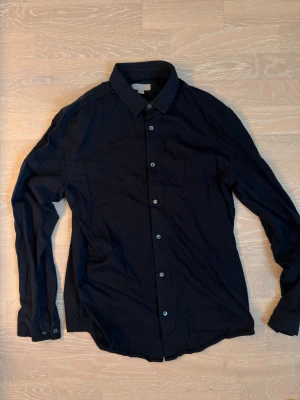 Mörkblå klassisk skjorta - Stilren mörkblå skjorta med lång ärm och knappar framtill. Skjortan har en klassisk krage och är tillverkad i ett mjukt tyg som känns bekvämt mot huden. Perfekt för dig som gillar en enkel och clean look.