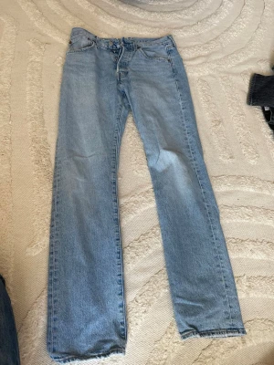Levis 501 - Säljer ett par ljusblå jeans med rak passform och klassisk femficksdesign. Jeansen har normal midja och stängs med dragkedja och knapp. Tillverkade i denim, perfekta för en avslappnad och trendig look.
