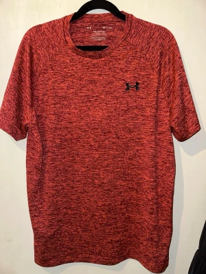 Röd tränings-tshirt från Under Armour - Snygg rödmelerad tränings-tshirt från Under Armour med svart logga på bröstet. Tillverkad i ett lätt och snabbtorkande funktionsmaterial som passar perfekt till gymmet eller löprundan. Klassisk rund halsringning och korta ärmar för maximal rörelsefrihet.