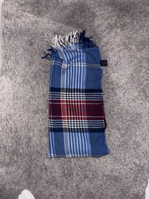 Blårutig halsduk från GANT - Snygg halsduk från GANT i mjukt material med klassiskt rutmönster i blått, vitt, svart och vinrött. Halsduken har fransar i ändarna och känns både stilren och tidlös. Perfekt accessoar för att lyfta din outfit under kyliga dagar.