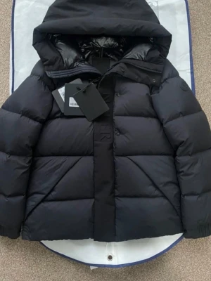 Moncler madeira  - Säljer en svart dunjacka från Moncler med stor huva och quiltad design. Jackan har en stilren look med diskret logotyp på ärmen och dragkedja samt knappar framtill. Perfekt för kalla vinterdagar, tillverkad i slitstarkt material och fylld med dun.
