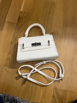 Vit liten handväska med axelrem - Snygg vit handväska i konstläder med stilren silverfärgad vridknäppe framtill. Väskan har ett topphandtag och en avtagbar, justerbar axelrem så du kan bära den på flera sätt. Perfekt till dig som gillar minimalistisk och trendig design.