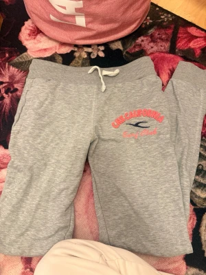 Gråa mjukisbyxor California Surf - Säljer ett par gråa mjukisbyxor med trycket 'California Surf Club' i rosa och blått på ena benet. Byxorna har bred resår i midjan och vit dragsko för skön passform. Perfekta för chill dagar eller när du vill vara extra bekväm.