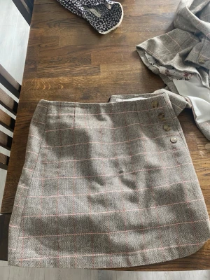 Rutig kortkjol och kavaj  - Säljer en snygg rutig kortkjol och kavaj från Tu Woman i beige och bruna toner med tunna röda ränder. Kjolen har knappar på sidan och är fodrad med vitt tyg. Perfekt för dig som gillar klassisk stil med en modern twist.