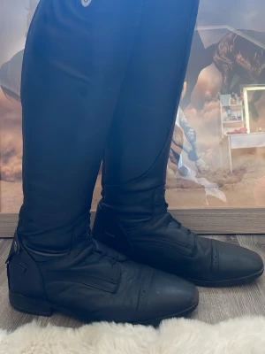 Svarta ridstövlar från CRW - Säljer ett par klassiska svarta ridstövlar från CRW i äkta läder. Stövlarna har hög modell med dragkedja bak och dekorativ knappdetalj upptill. Perfekta för dig som vill ha en stilren och funktionell look i stallet. Storlek 40. Pris 200kr