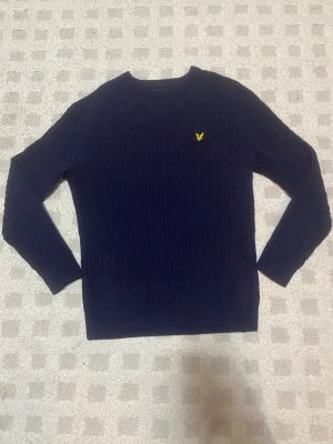 Mörkblå stickad tröja Lyle & Scott - Mörkblå stickad tröja från Lyle & Scott med klassisk gul broderad örn på bröstet. Tröjan har rund halsringning, ribbstickade muddar och ett diskret ribbat mönster i materialet. Perfekt för dig som gillar stilrena och tidlösa plagg. 